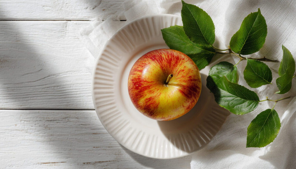 Top view of red-yellow apple on ceramic plate with green leaves on white rustic background