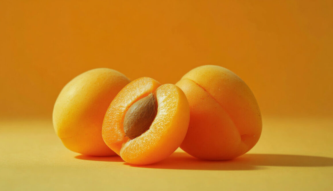Three fresh apricots on vibrant orange background, one sliced to show pit, photographed in sharp 16:9 format