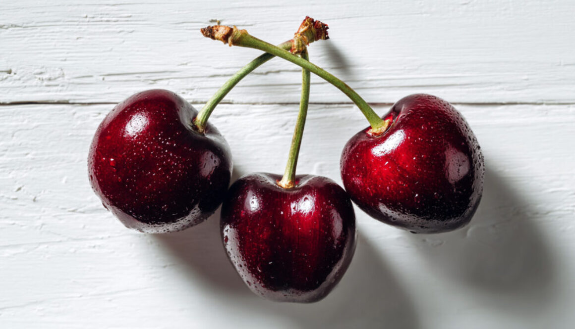Flat lay of three fresh cherries with water droplets on a rustic white wooden background, realistic fruit photo in 16:9 format