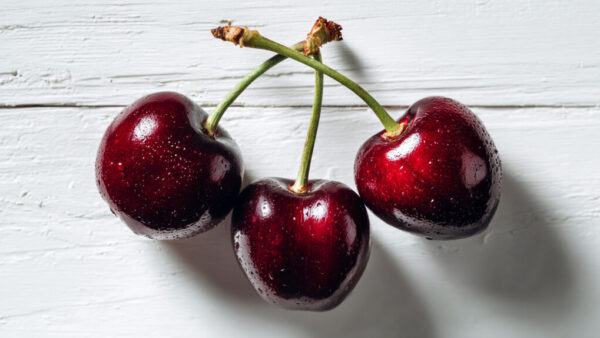 Flat lay of three fresh cherries with water droplets on a rustic white wooden background, realistic fruit photo in 16:9 format