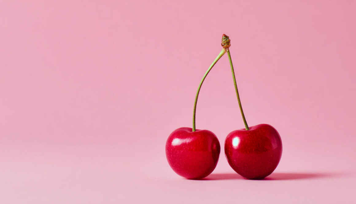 Two red cherries with crossed stems centered on pink solid background in 16:9 format
