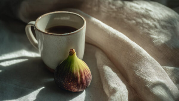 Fresh fig placed next to a ceramic coffee cup on soft linen cloth with natural light in 16:9 format