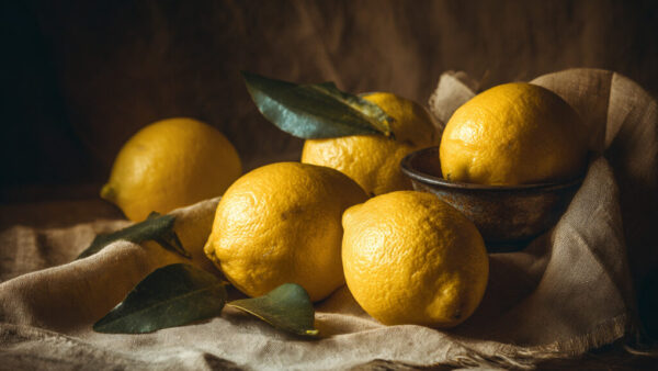 Rustic still life of fresh lemons with leaves on fabric and ceramic bowl, dark background