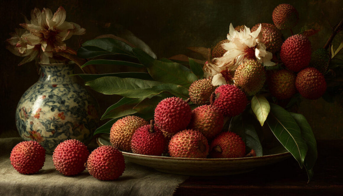 Artistic still life of lychees, leaves, and flowers beside a ceramic vase on dark background in 16:9 format